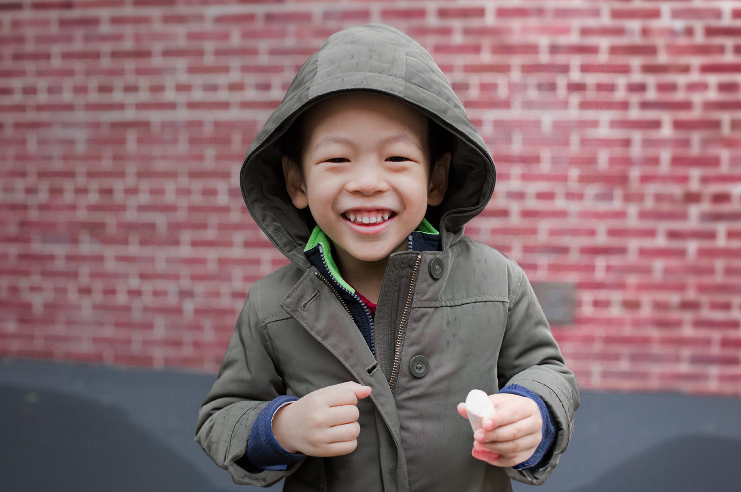 Portrait of a child with his hood up smiling in front of a brick wall holding sidewalk chalk